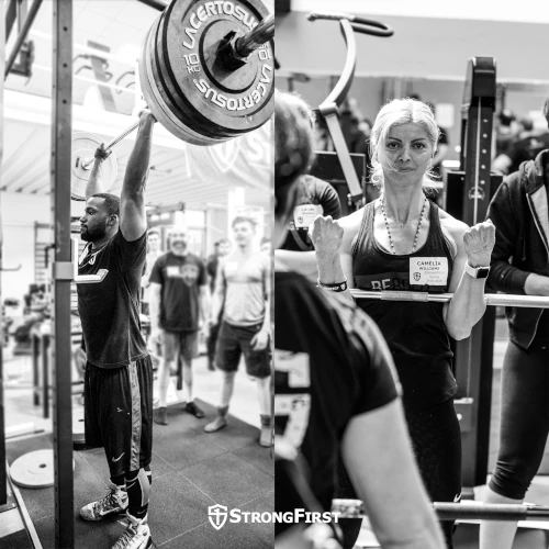 A man does a barbell military press while a woman stands at the top of a Zercher squat.